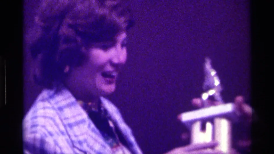Woman in a blue plaid jacket smiles or laughs while a gold-colored trophy is handed toward her in a Kentucky 1976 interior.