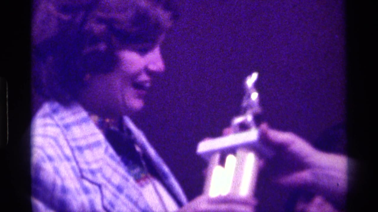 Woman in patterned blazer smiles as a trophy is handed toward her on a dim stage.