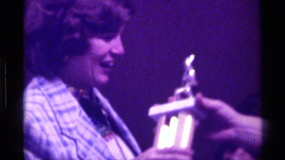 In 1976 Kentucky 8mm footage, a woman in a patterned blazer smiles while receiving a small trophy under purple stage light.