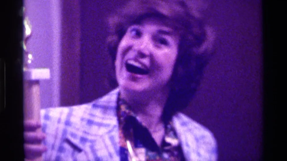 The woman laughs broadly while holding the trophy, with a dark backdrop and purple lighting visible in the 1976 Kentucky home movie frame.