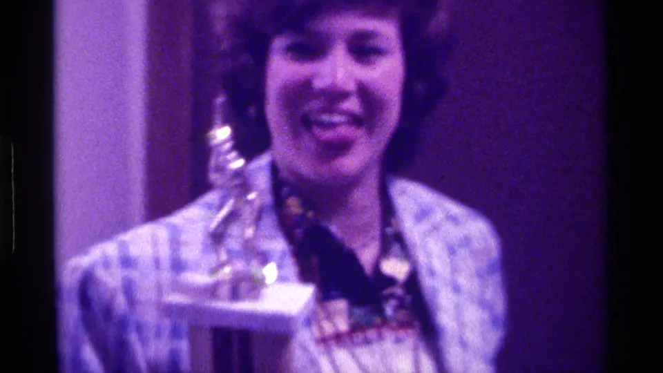 The woman smiles with the trophy held near her chest, framed closely against a dark purple-lit backdrop.