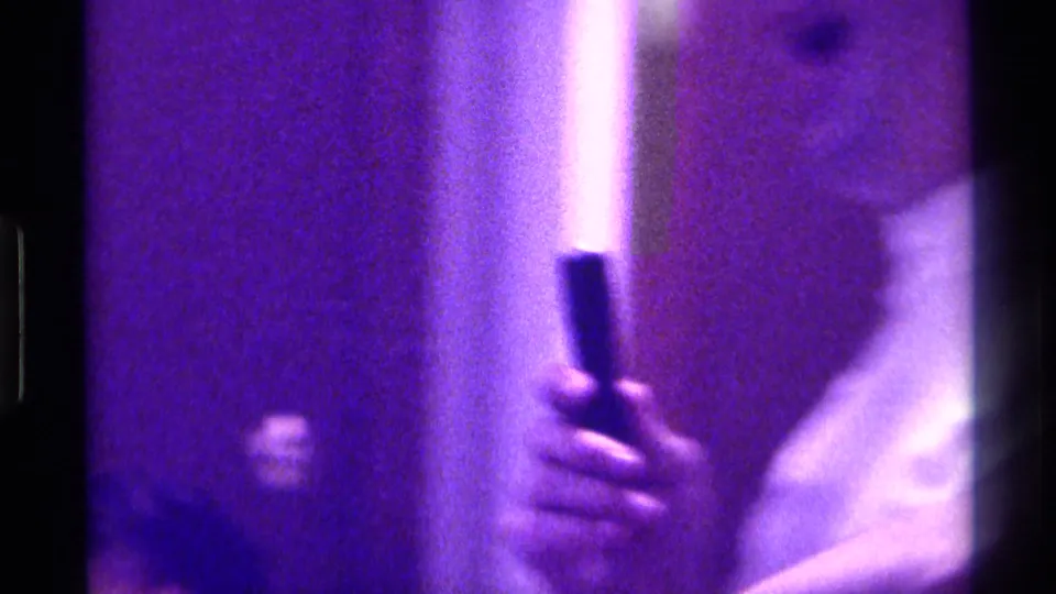 Close 1970s home-movie view of a handheld microphone held near a person in a light shirt, with heavy purple cast.