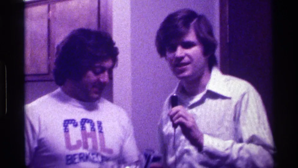1976 Kentucky home-movie frame of two young men indoors, with one wearing a CAL Berkeley T-shirt and the other holding a microphone.