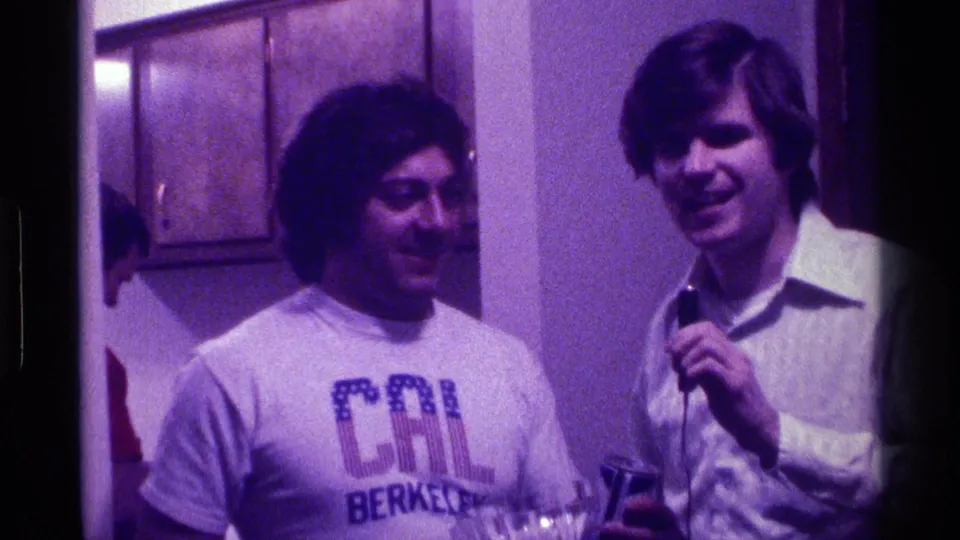 1976 Kentucky home-movie frame showing two young men smiling indoors, one in a CAL Berkeley T-shirt and one holding a microphone.