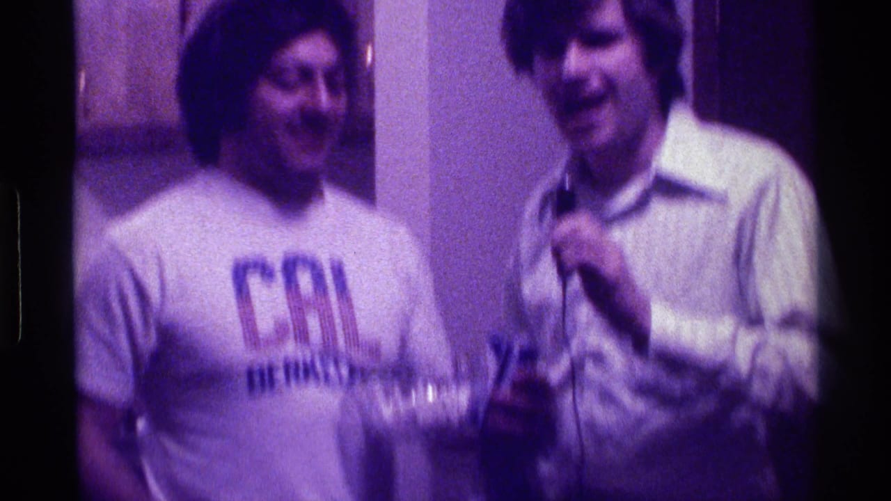 Two adult men stand indoors, one wearing a Cal Berkeley T-shirt and one holding a microphone.