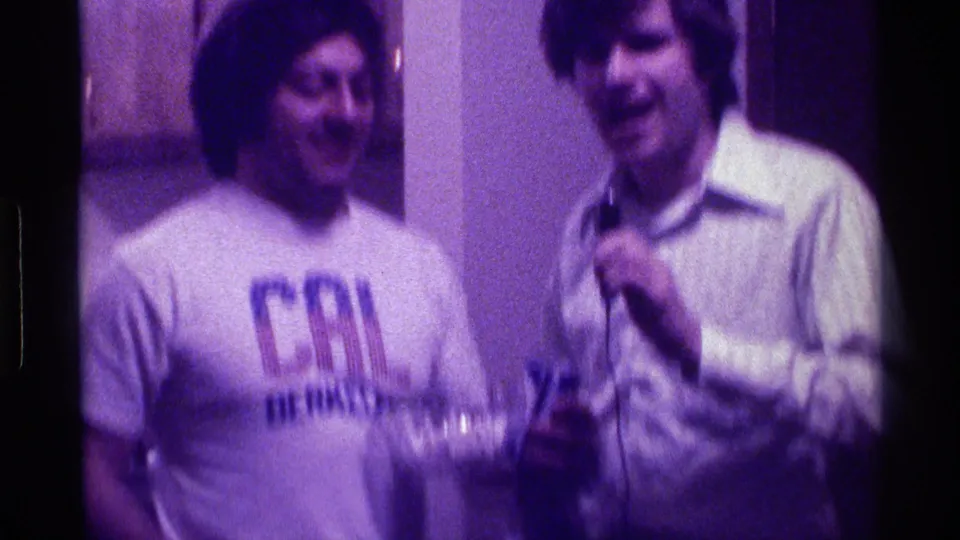 In a 1976 Kentucky home-movie frame, two adult men stand at an indoor party as one smiles and the other holds a microphone.