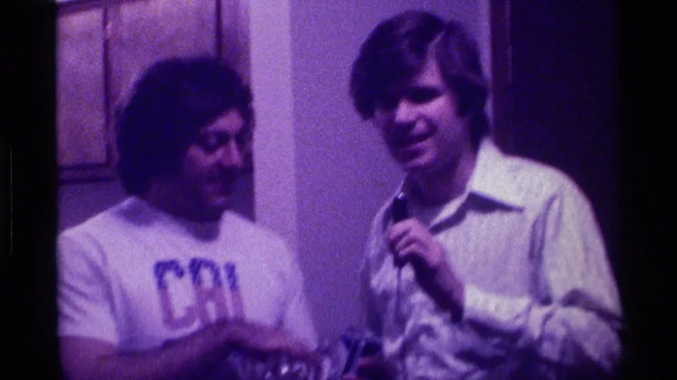 The man in the Cal Berkeley T-shirt looks toward the microphone holder in a purple-tinted 1976 Kentucky indoor party frame.