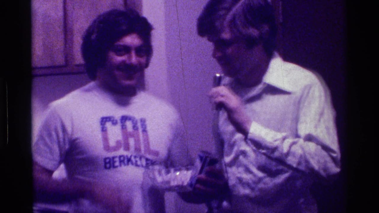 1976 Kentucky home-movie frame of two young men indoors, with a CAL Berkeley T-shirt, handheld microphone, bowl, and soda can visible.