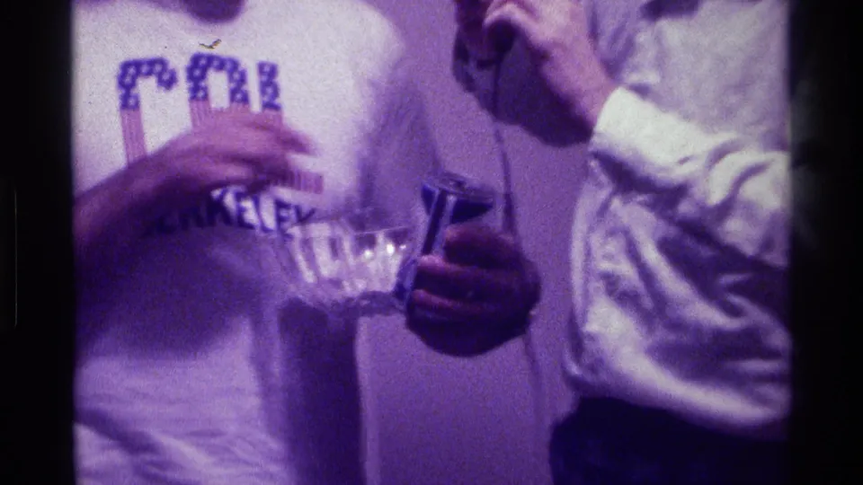 Close 1976 Kentucky home-movie frame showing a CAL Berkeley shirt, glass bowl, soda can, and microphone during an informal indoor exchange.