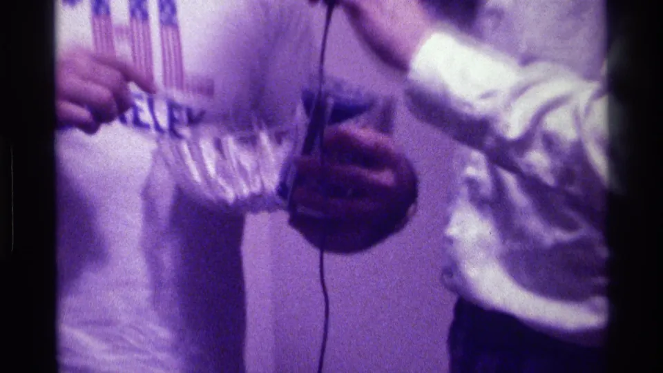 1976 Kentucky home-movie close-up of hands holding a glass bowl near a soda can and microphone, with the CAL Berkeley shirt partly visible.