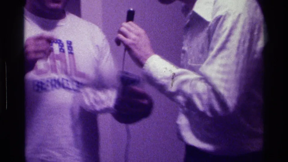 1976 Kentucky home-movie frame of one young man gesturing beside another holding a microphone and soda can in a private interior.