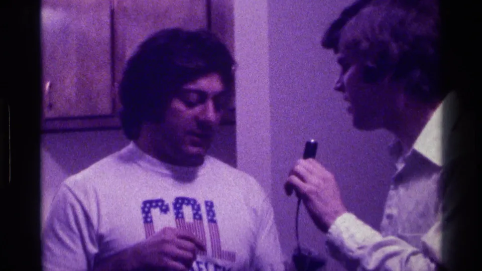 1976 Kentucky home-movie frame of two young men indoors, with one holding a small microphone near a man in a CAL Berkeley shirt.
