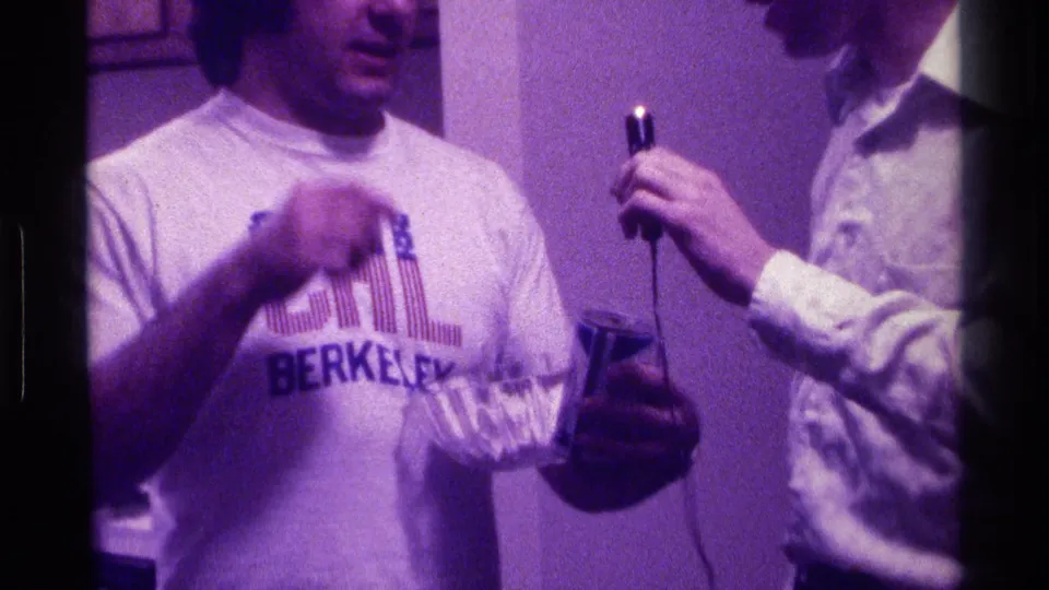 1976 Kentucky party frame showing a man in a CAL Berkeley shirt gesturing beside a glass bowl while another man holds a microphone.