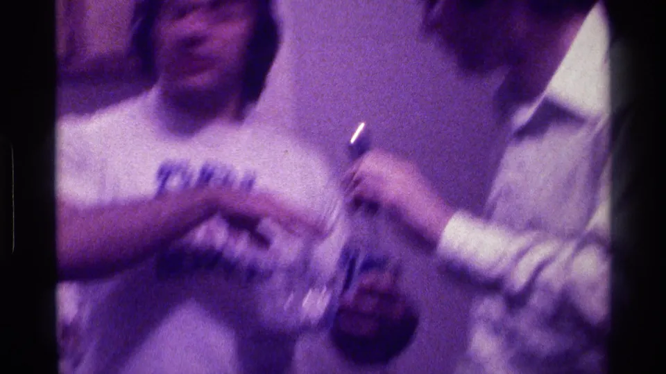 Soft, motion-blurred 1976 Kentucky home-movie frame of two men during a microphone game or party exchange indoors.