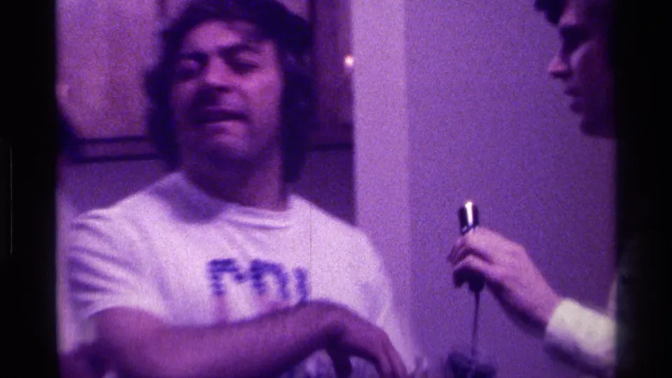 1976 Kentucky frame of a man in a CAL Berkeley shirt beside a microphone, with a second young man partly visible at right.