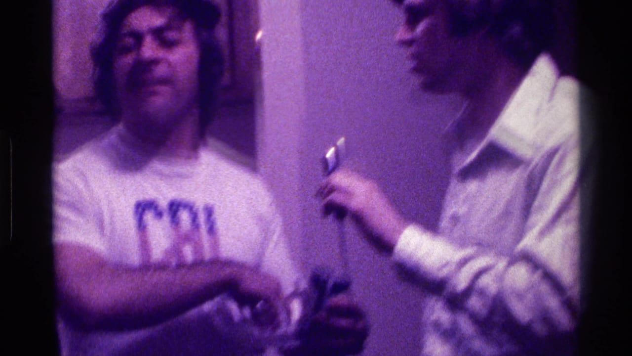 Two adult men stand indoors as one holds a small microphone near the other.