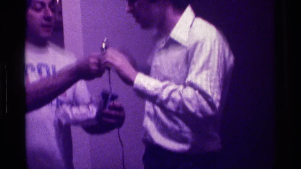 The man in the light button-down holds a small wired microphone as the man in the CAL shirt reaches toward it in a private interior.