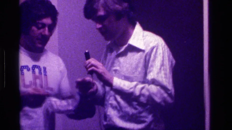 The two men stand close together in the purple-tinted 8mm frame, looking down as the microphone is handled between them.