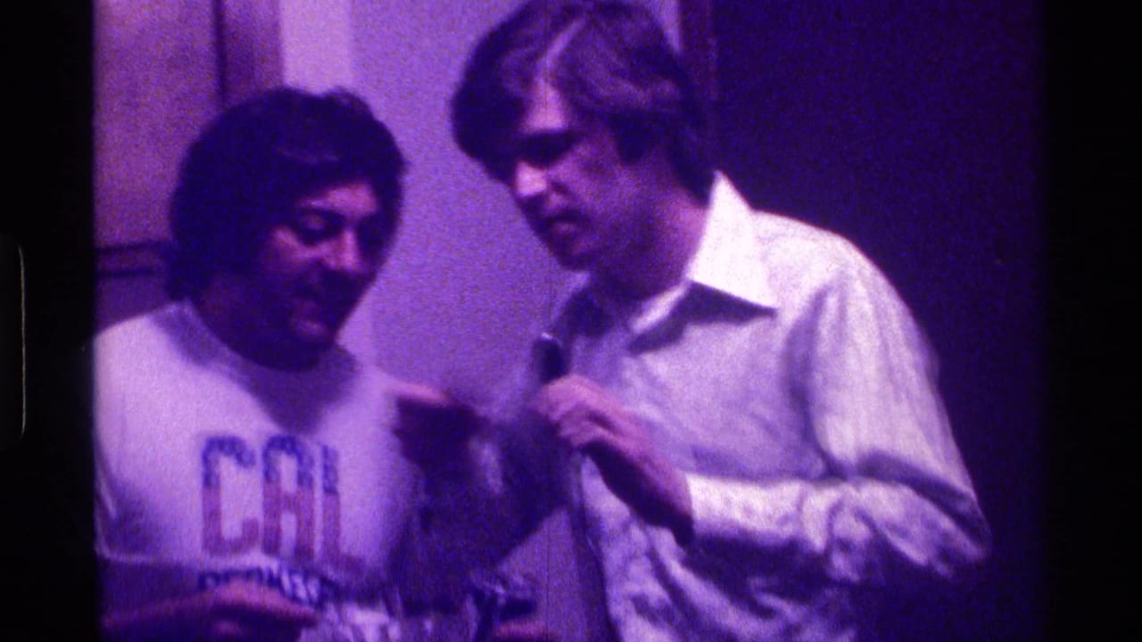 Two young men stand close together indoors, one holding a microphone under purple light.