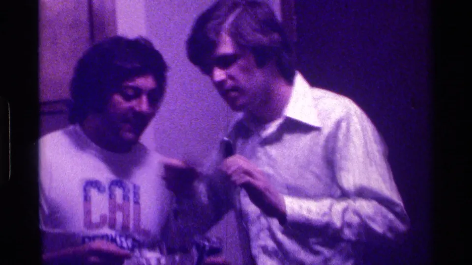 In 1976 Kentucky home-movie footage, two young men stand in a dim room as one holds a microphone and the other wears a printed T-shirt.
