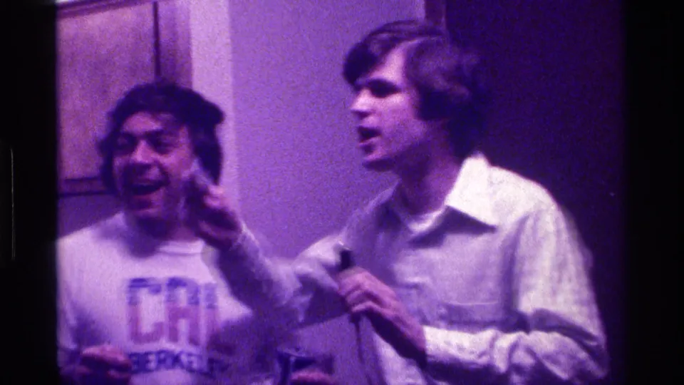 A young man in a pale shirt gestures with one hand while holding a microphone beside a smiling friend in a purple-lit room.
