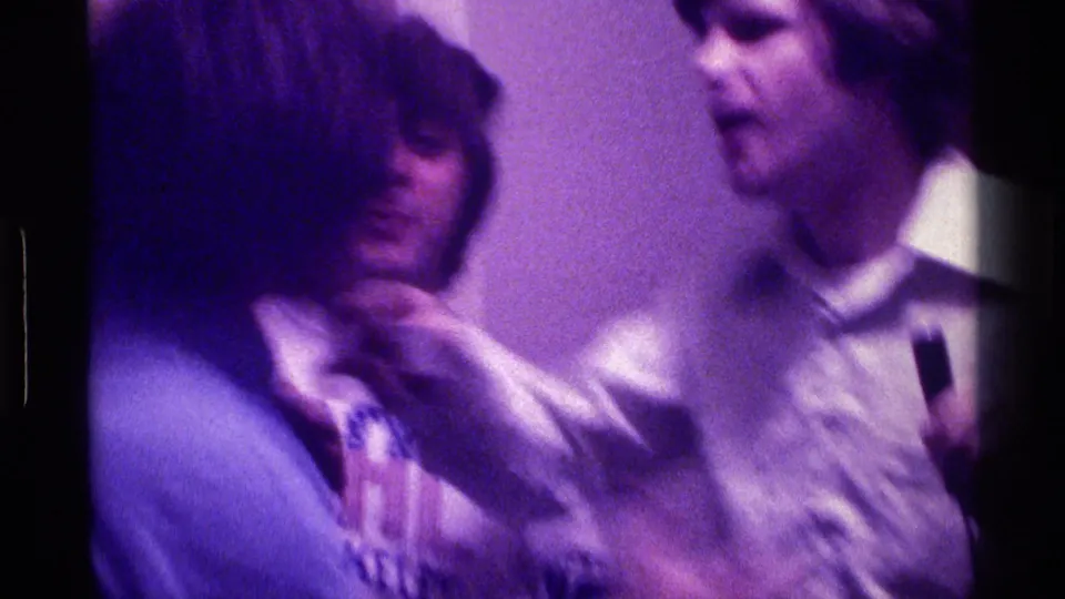 Inside a dim Kentucky room, a young man with longer hair appears to speak as a friend holds a microphone nearby in soft 8mm focus.