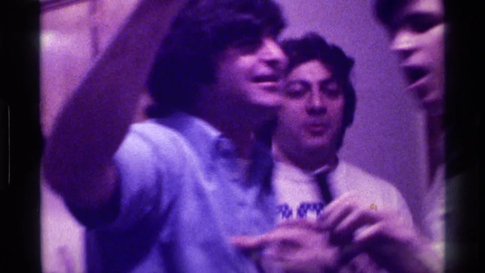 Several young adults crowd together in a purple-lit interior, with one raised arm near the lens and a printed T-shirt visible in the background.
