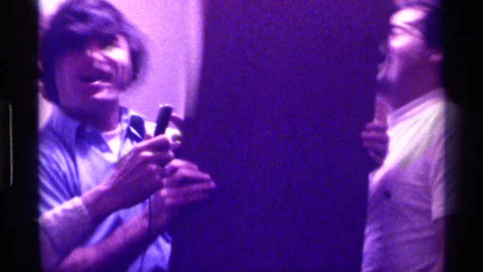 A young man holds a microphone while another stands nearby in purple-tinted 1976 Kentucky home-movie footage.