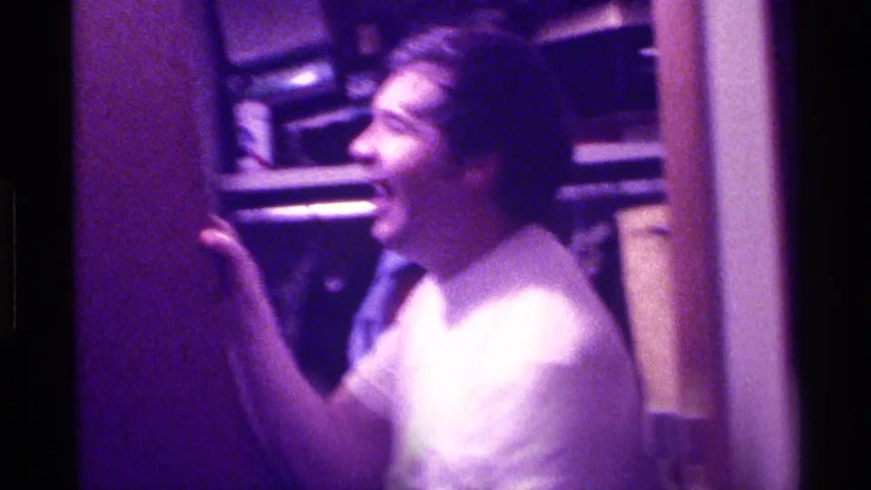 A young man smiles beside shelves or books in a purple-lit private Kentucky interior from 1976 home-movie footage.