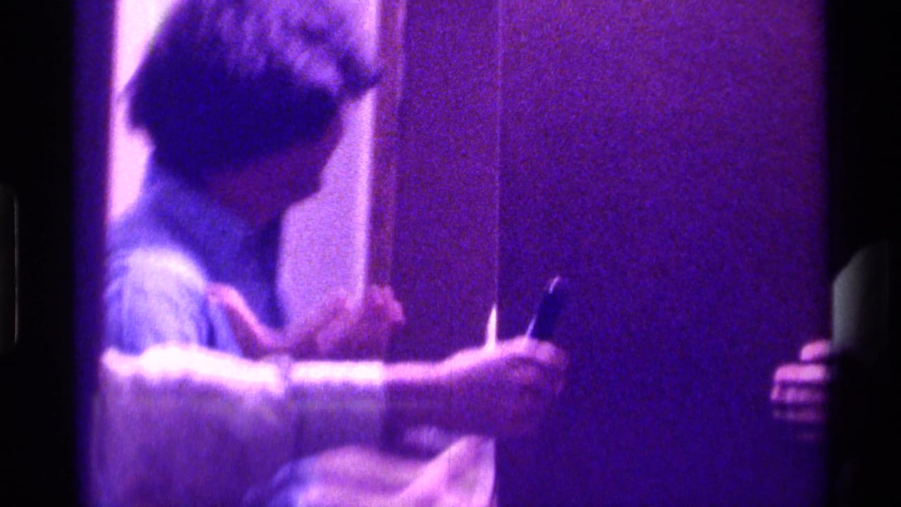 In 1976 Kentucky home-movie footage, a young man in a light shirt stands near a doorway in a dim interior with a strong purple cast.