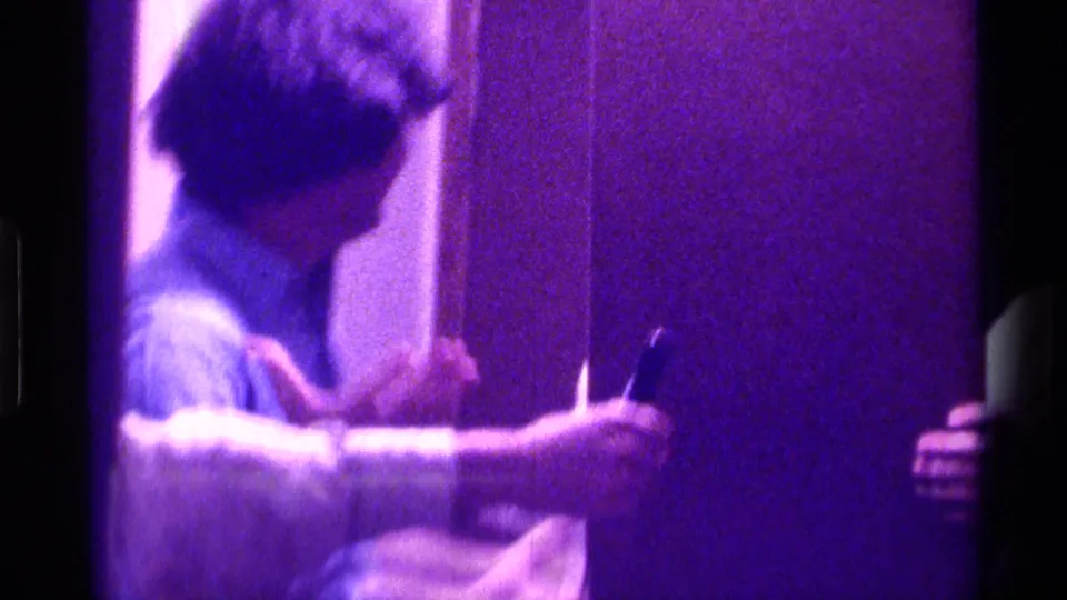 In 1976 Kentucky home-movie footage, a young man in a light shirt stands near a doorway in a dim interior with a strong purple cast.
