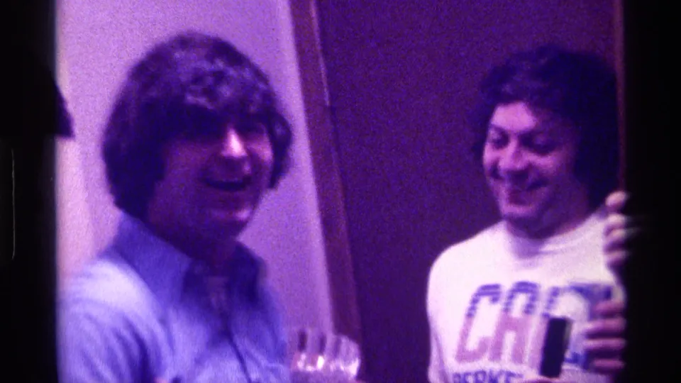 Two young men smile and face the camera inside a dim Kentucky party interior, with one shirt showing visible lettering.
