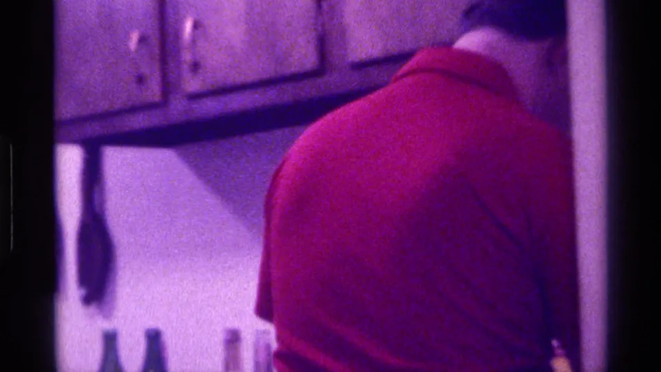 A person in a red shirt is seen from behind in a private interior with wood cabinets and dim Super 8 color.