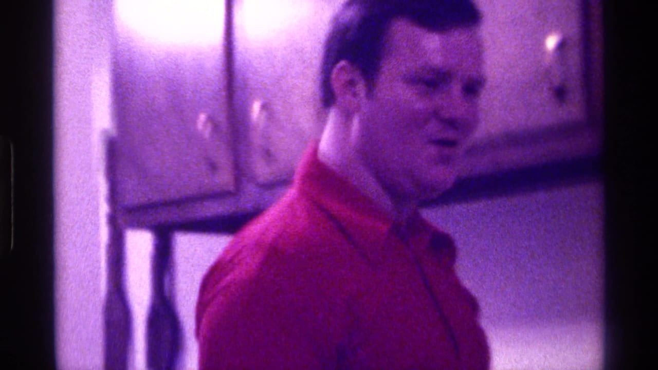 In 1976 Kentucky home-movie footage, an adult man in a red collared shirt stands in a kitchen-like room with wooden wall cabinets.