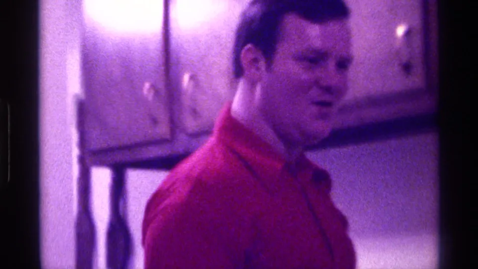 In 1976 Kentucky home-movie footage, an adult man in a red collared shirt stands in a kitchen-like room with wooden wall cabinets.