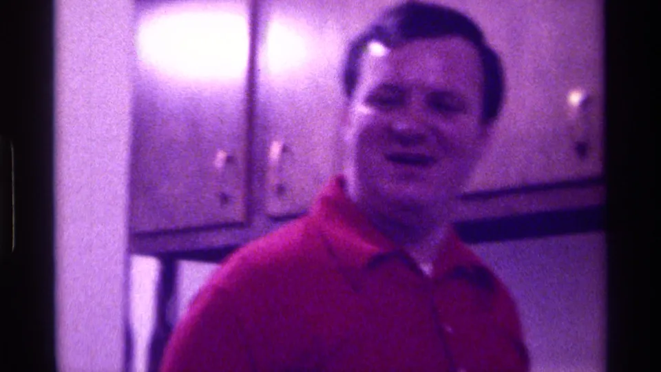 The man in the red shirt faces the camera and appears to smile in a 1976 Kentucky private interior with wall cabinets behind him.