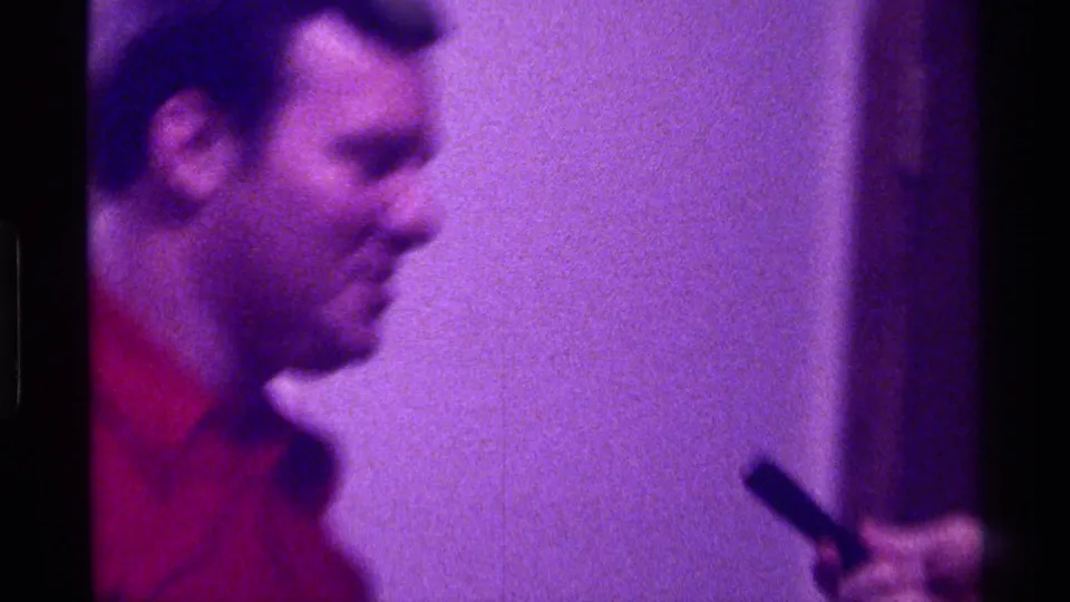 A close, soft-focus profile view shows the man in the red shirt speaking or gesturing with a small dark object inside the room.