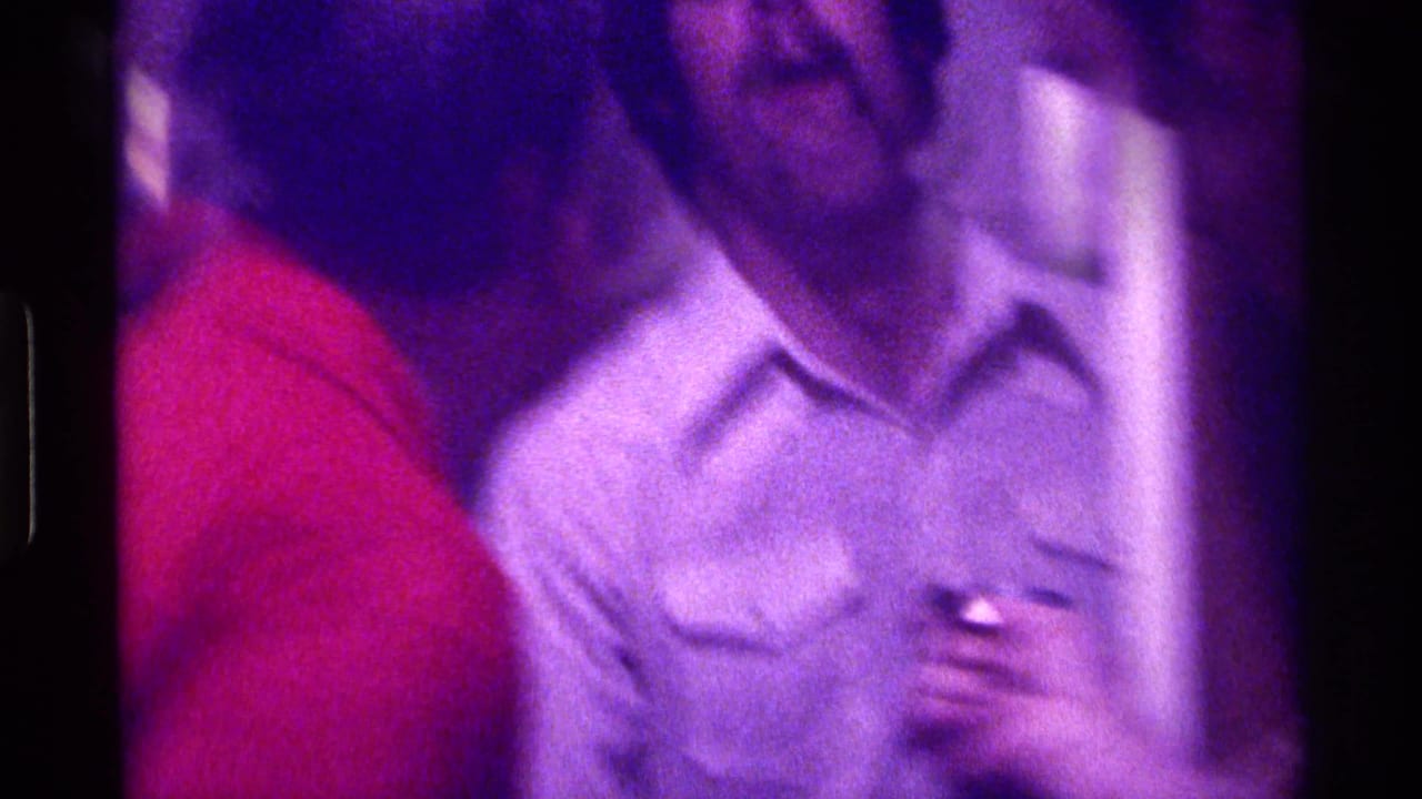 Two adult men stand close indoors, one in a red shirt and one in a light button-up shirt.
