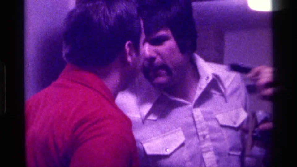 1976 Kentucky indoor home-movie frame with two men in close conversation and a microphone extended toward the mustached man.