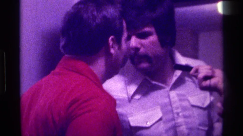 Purple-tinted 1976 Kentucky 8mm frame showing a red-shirted man facing a mustached man with a microphone near his shoulder.