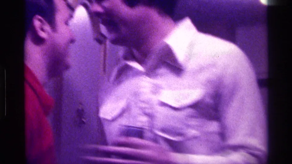 1976 Kentucky home-movie frame of a man in a light button-up leaning toward another man indoors, with strong motion blur and magenta cast.