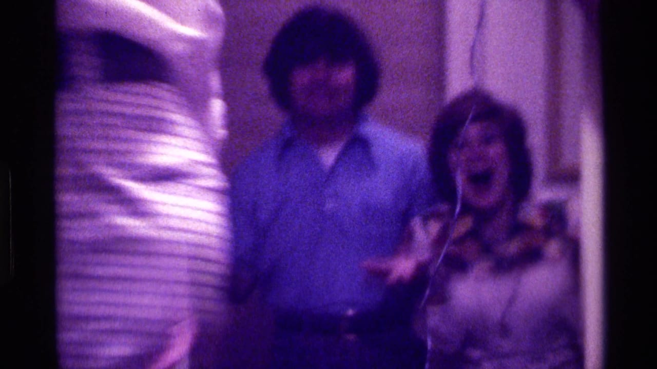 Three adults in a dim interior, with a man in blue and a woman in a patterned top laughing.