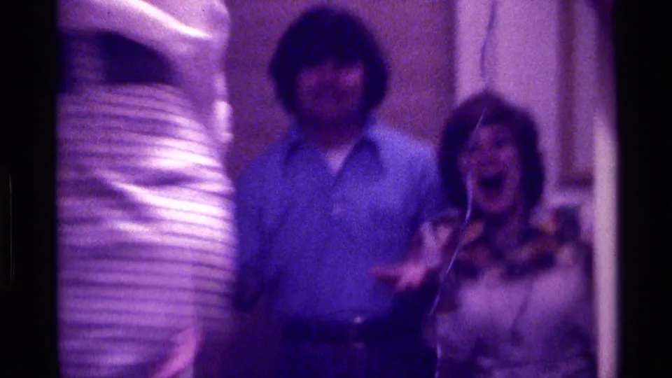 1976 Kentucky 8mm frame of adults in a private room, including a man in blue and a woman in a patterned top reacting to the camera.