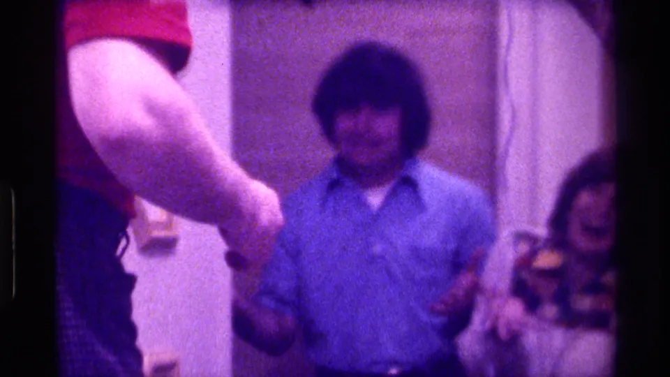 1976 Kentucky home-movie frame showing a man in blue near center, a red shirt sleeve at left and a laughing woman partly visible at right.