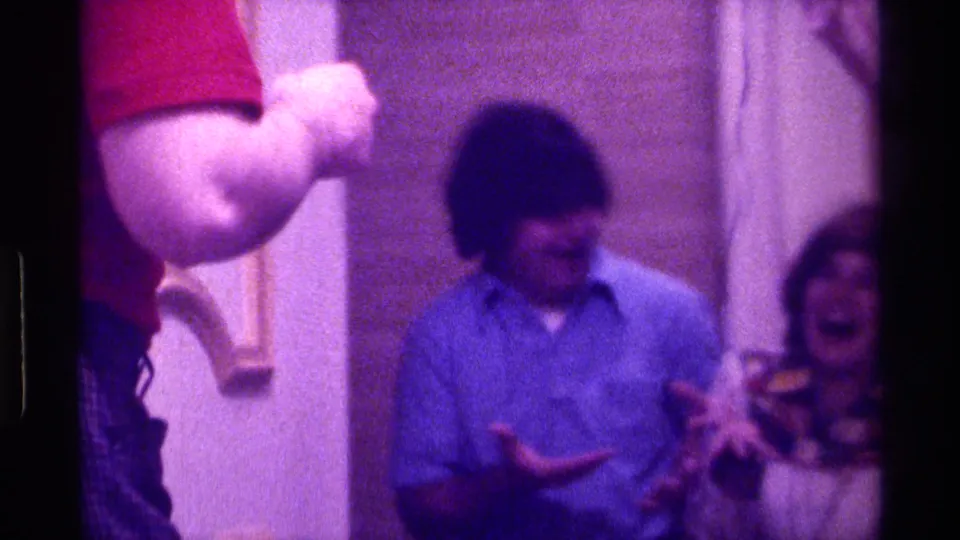 Soft 1976 Kentucky 8mm frame with a red-shirted adult at left, a man in blue looking down and a laughing woman partly visible behind him.
