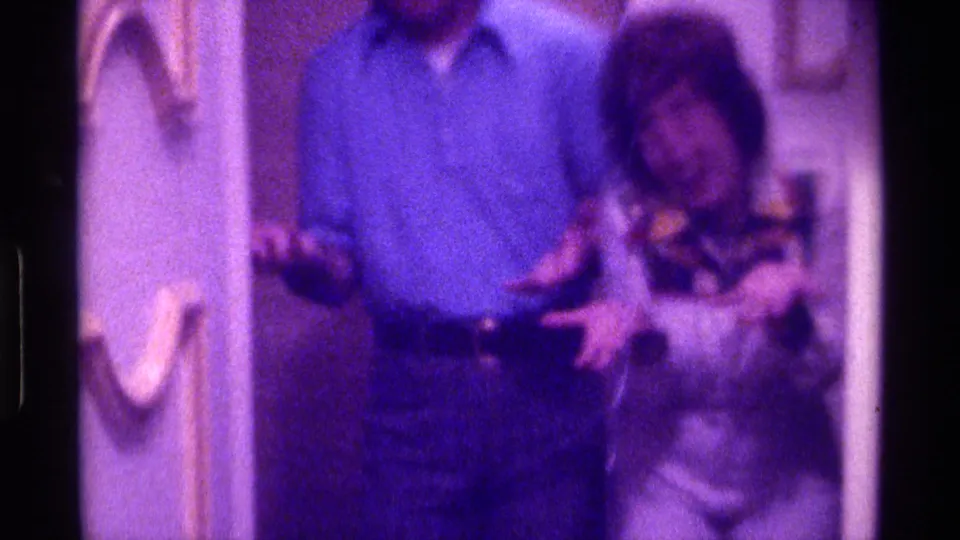 1976 Kentucky home-movie frame of a man in blue and a woman in a patterned top standing close in a softly lit private interior.