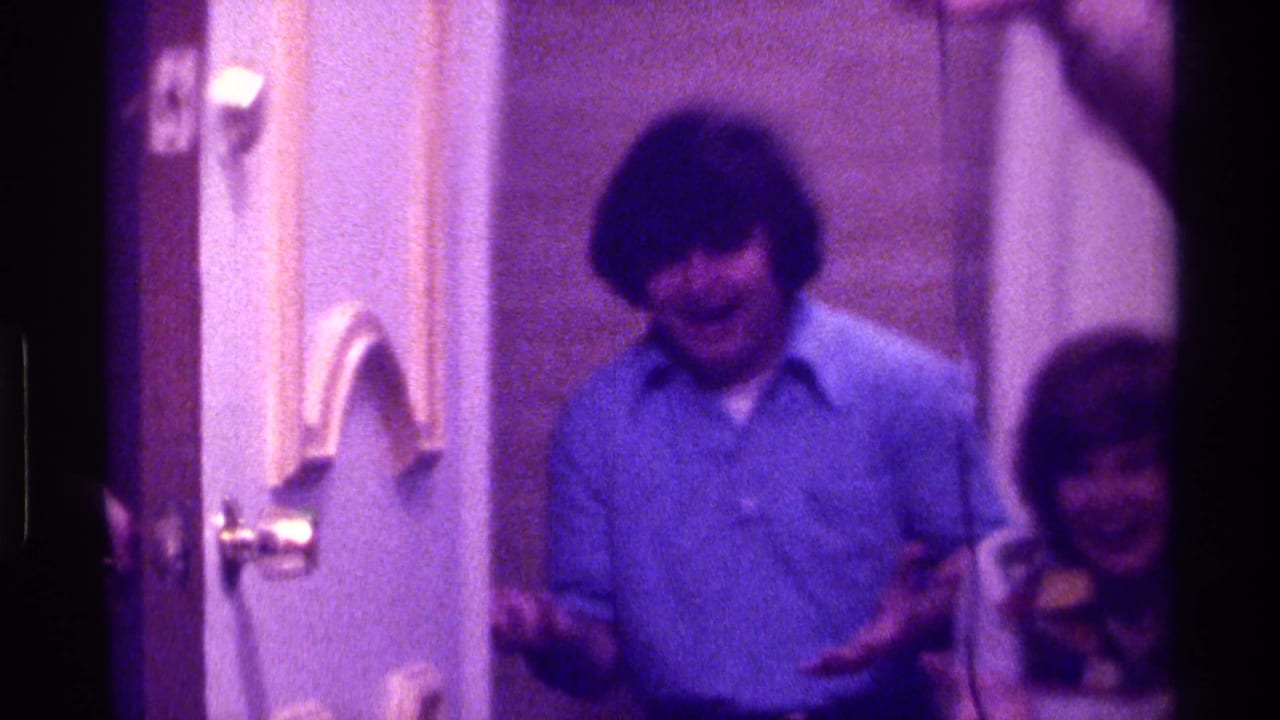 Two adults stand near an interior doorway as one man smiles toward the camera.