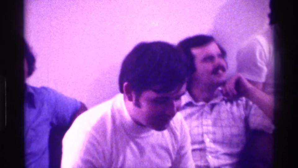 Men sit close together in a Kentucky home interior, with a white T-shirt in the foreground and a mustached man in a collared shirt behind him.