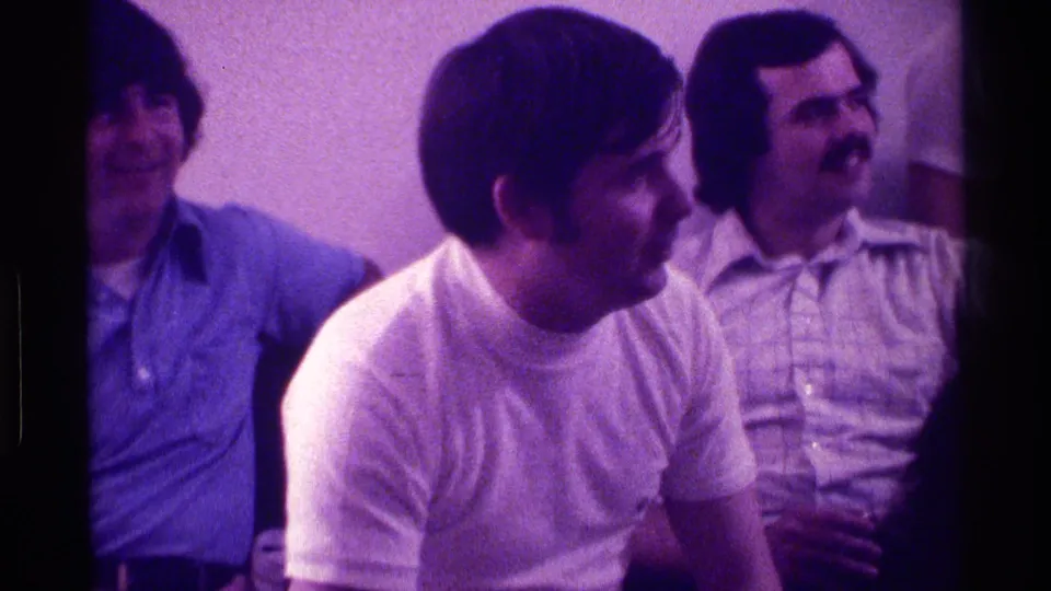 A group of men sit in a Kentucky interior, looking off-camera while one mustached man holds a glass during the gathering.