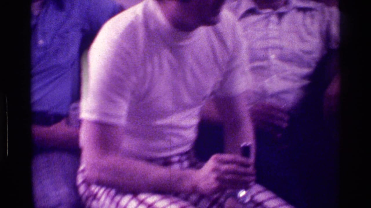 Seated men in casual shirts, one in a white t-shirt leaning forward while holding a drink.